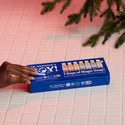 A hand reaches for a blue box labeled Dr. Bronners 6 Pack of Magic Soaps on a tiled surface, surrounded by green branches. The package features various Dr. Bronners soap samples [One Size]