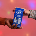 Two hands exchange a blue box labeled Dr. Bronners Joy Peppermint Essentials Gift Set against a pink background with colorful lights. The packaging promotes Dr. Bronners pure castile soap products [One Size]