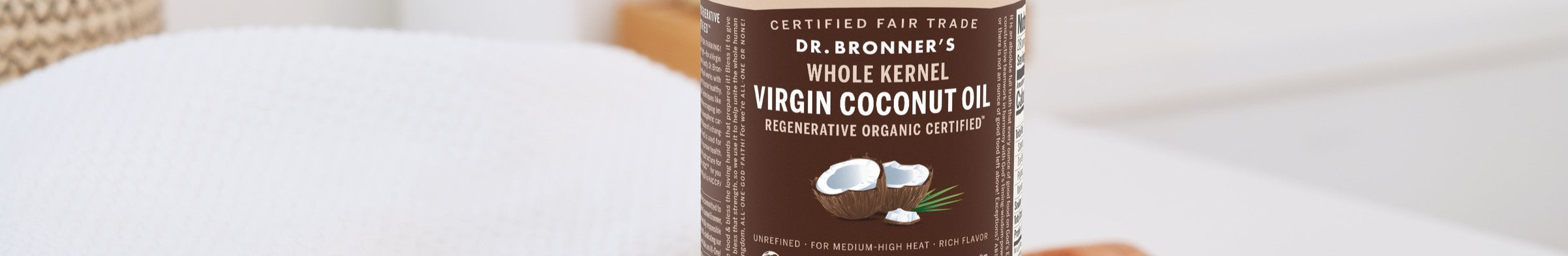 Dr. Bronners Virgin Coconut Oil jar sits on a textured white surface, featuring text CERTIFIED FAIR TRADE, WHOLE KERNEL, and REGENERATIVE ORGANIC CERTIFIED, with coconut illustrations. Its in a minimalist room setting