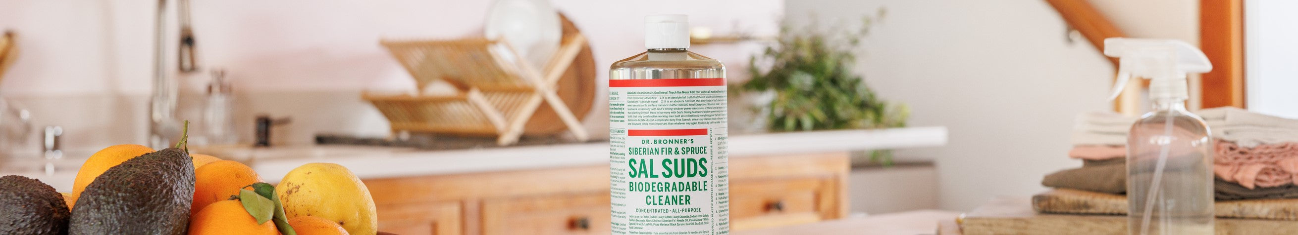 A bottle of Dr. Bronners Sal Suds Biodegradable Cleaner sits on a kitchen countertop. Nearby are fresh fruits, folded towels, and a spray bottle, emphasizing an eco friendly cleaning environment
