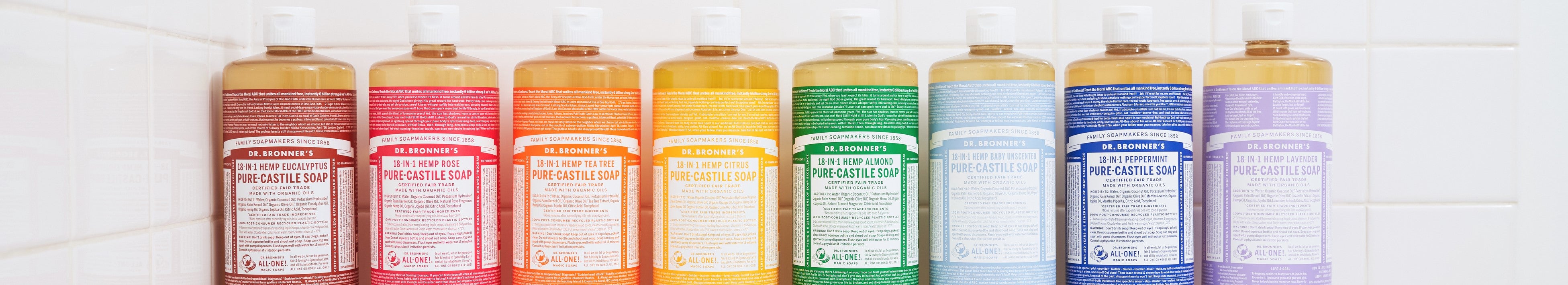 Bottles of Dr. Bronners Pure Castile Soap, in various scents like eucalyptus, rose, tea tree, citrus, almond, unscented, peppermint, and lavender, line a tiled bathroom shelf, showcasing eco friendly packaging
