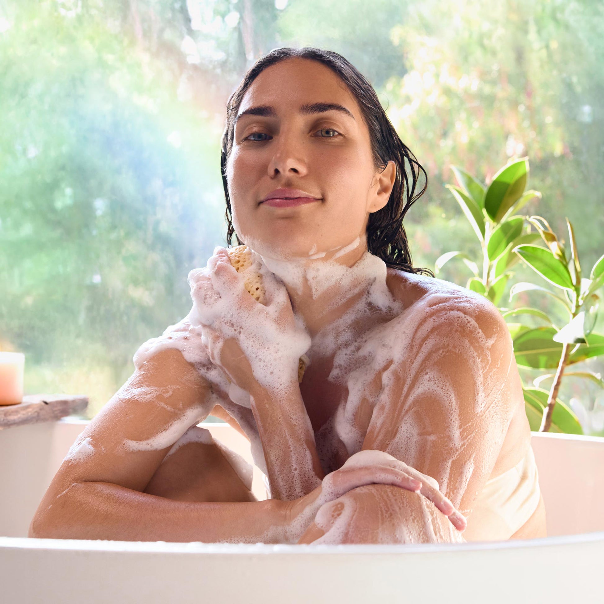 A person enjoys a relaxing bath, covered in frothy suds from soap, while sitting in a tub. Nearby, a plant and a candle enhance the serene, natural atmosphere [2 oz, 3.4 oz, 4 oz, 8 oz, 16 oz, 32 oz, 1/2 Gallon, 1 Gallon]