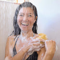 A person showers, smiling while lathering with a sponge. Water cascades from above, creating a refreshing atmosphere. The setting suggests a personal care routine using natural body wash [32 oz]