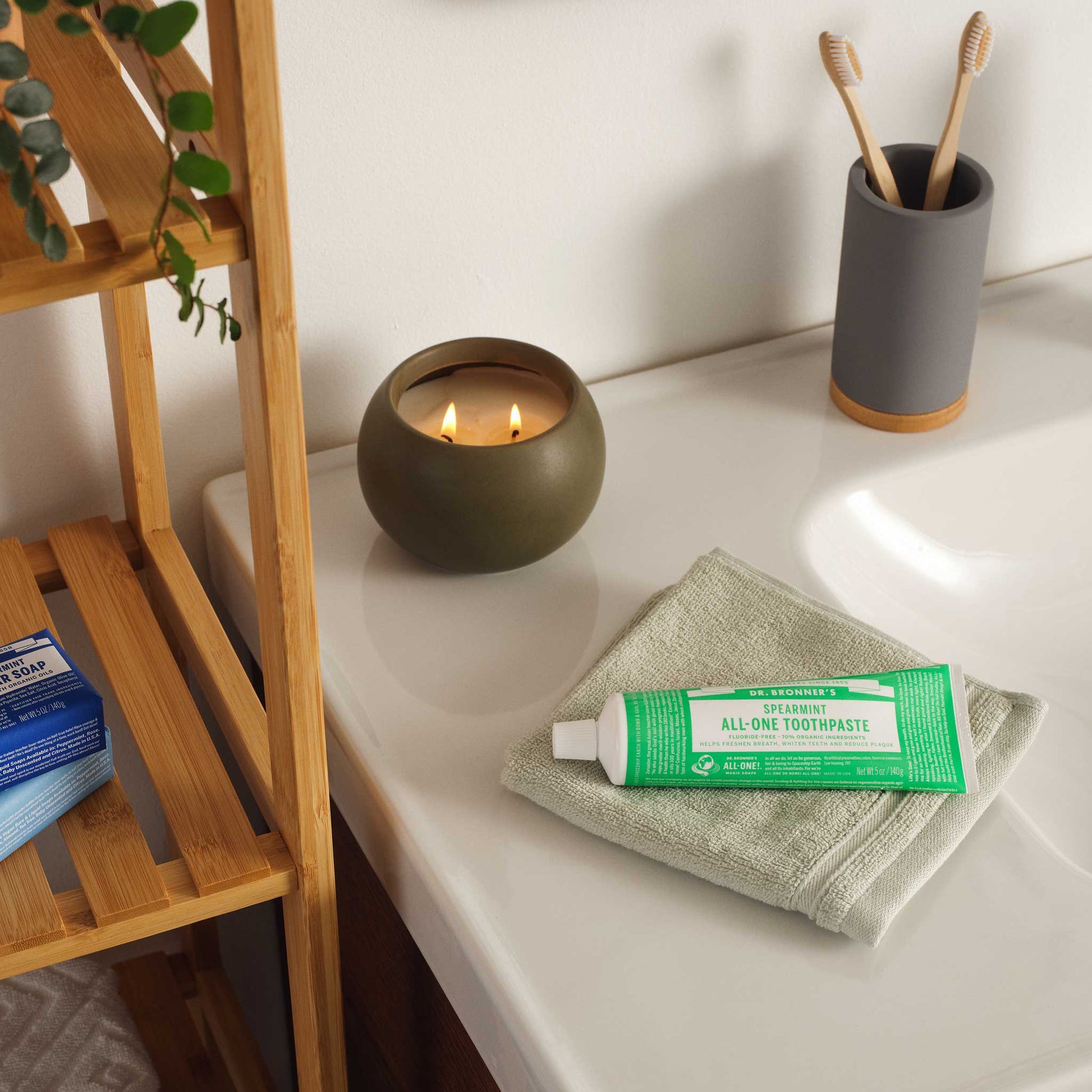 A green tube of toothpaste rests on folded towels beside a sink, with a candle and toothbrushes nearby, creating a serene and eco friendly bathroom setting [5 oz]