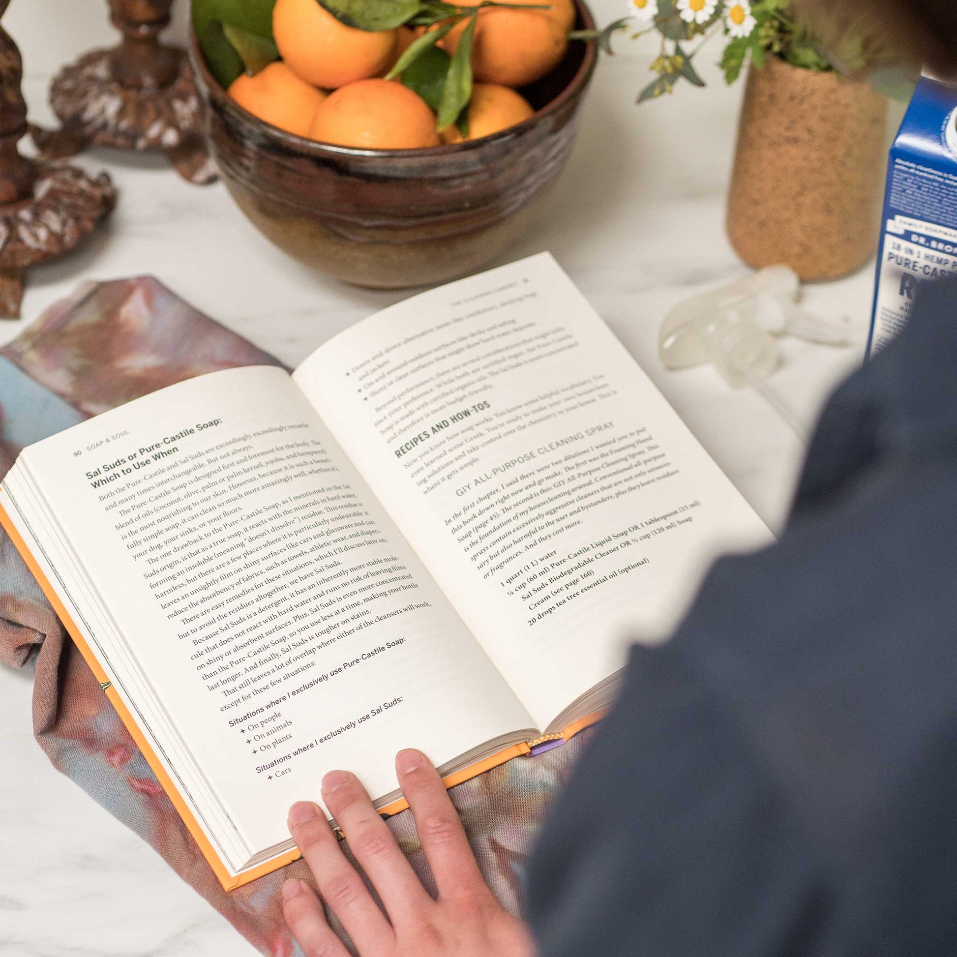 An open book displays a recipe for using Dr. Bronners Pure Castile Soap, with a persons hand holding the pages. Nearby, a bowl of oranges and flowers adds a fresh ambiance