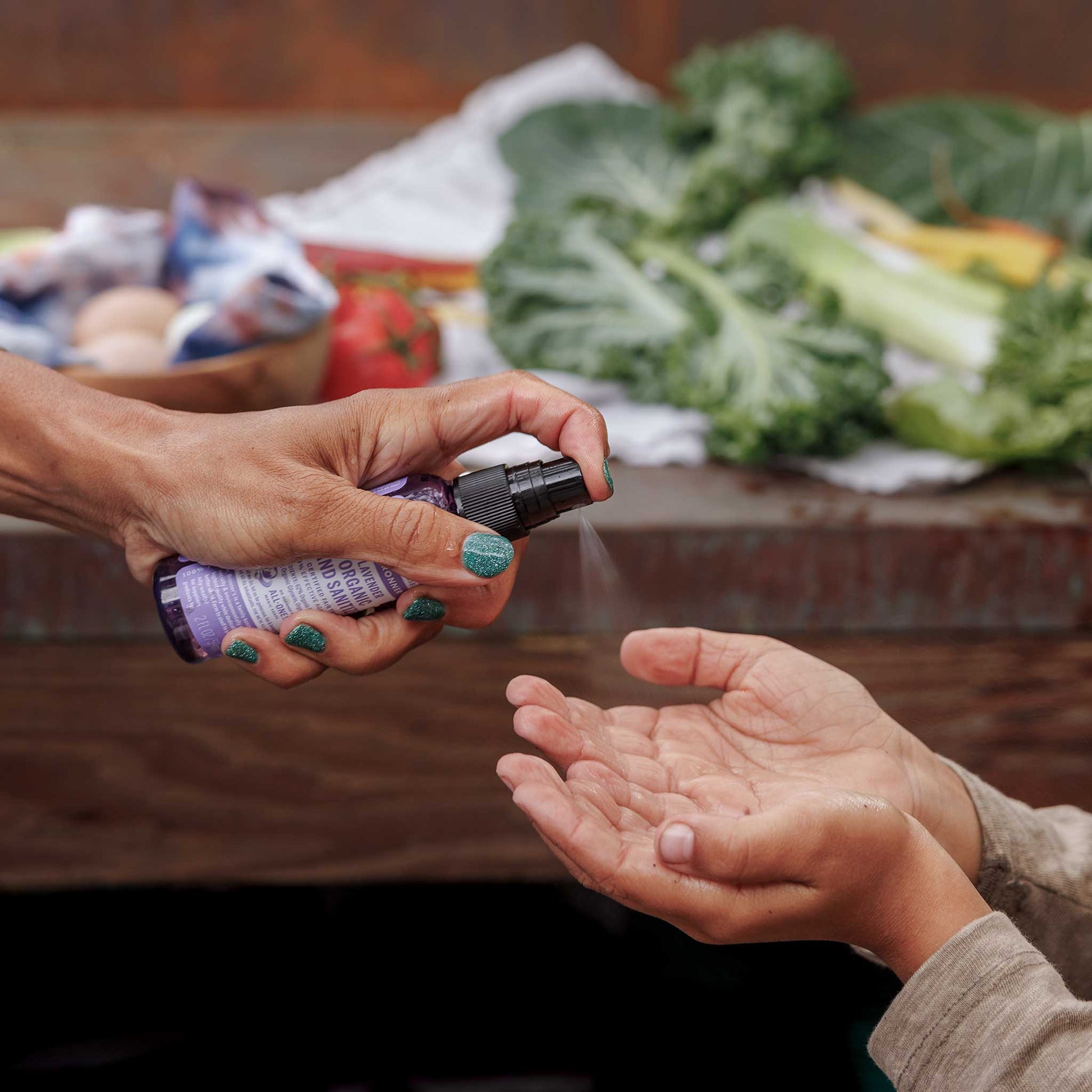 A hand dispenses liquid from a small pump bottle into another persons open palms, set against a background of fresh vegetables on a wooden surface, suggesting hygiene before food handling