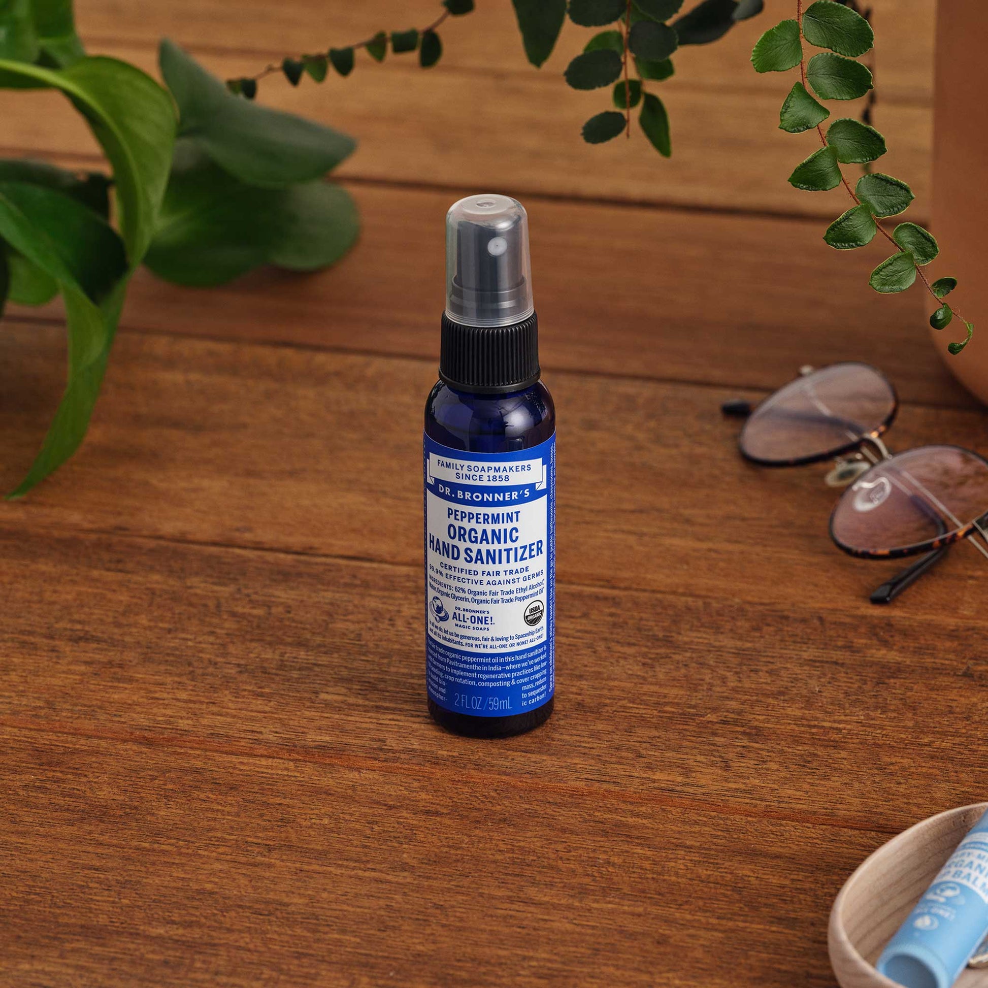 A 2 oz. bottle of Dr. Bronners Peppermint Organic Hand Sanitizer stands on a wooden surface near green leaves and sunglasses, emphasizing natural, eco friendly hygiene. Dr. Bronners Peppermint Organic Hand Sanitizer