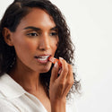 A person applies lip balm to their lips, holding a tube against a white background. They have curly hair and wear a white shirt, focusing on moisturizing their lips naturally [.15 oz]