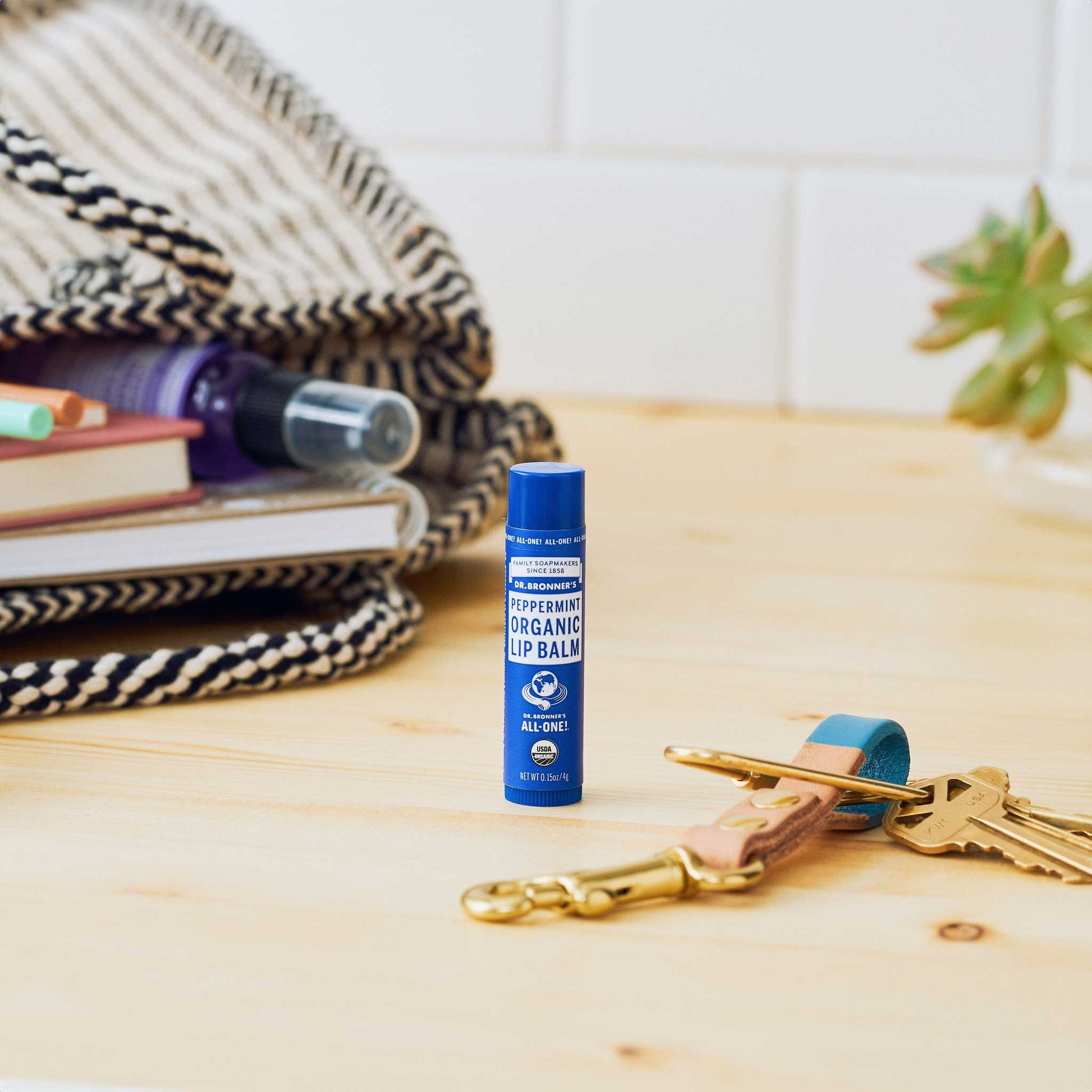 Dr. Bronners Peppermint Organic Lip Balm stands upright on a wooden table, surrounded by a keychain, books, and a woven bag in a cozy setting. Dr. Bronners Peppermint Organic Lip Balm. ALL ONE [.15 oz]