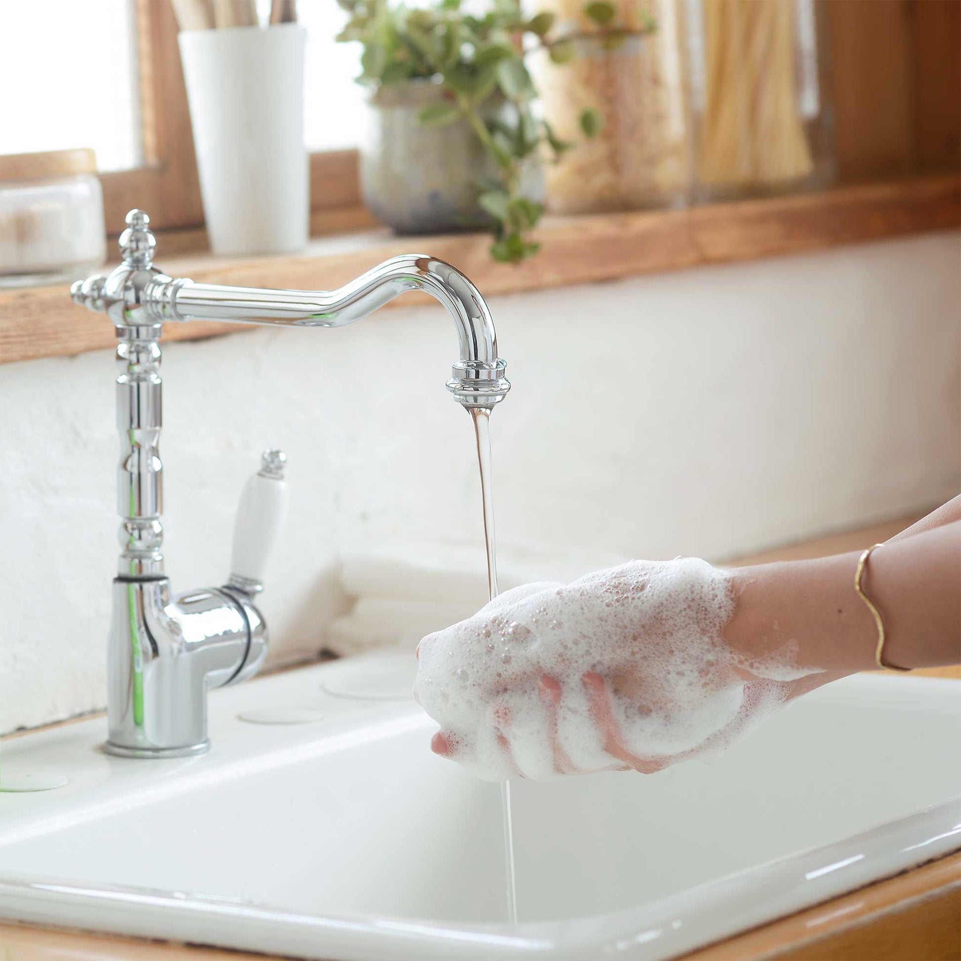 A hand lathers with foamy soap under a running faucet in a kitchen sink, surrounded by wooden counter accents and a potted plant for a fresh, natural ambiance [12 oz, 24 oz, 1/2 Gallon]