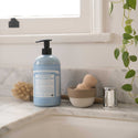 A bottle of Dr. Bronners liquid soap stands on a marble countertop beside grooming tools. The sunlit window and green plant enhance an eco friendly bathroom ambiance. Text on the bottle includes product details [24 oz]