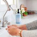 Hands lather with Dr. Bronners Organic Sugar Soap under a kitchen faucet, set against a bright countertop with decorative elements like flowers and dishes nearby [24 oz]