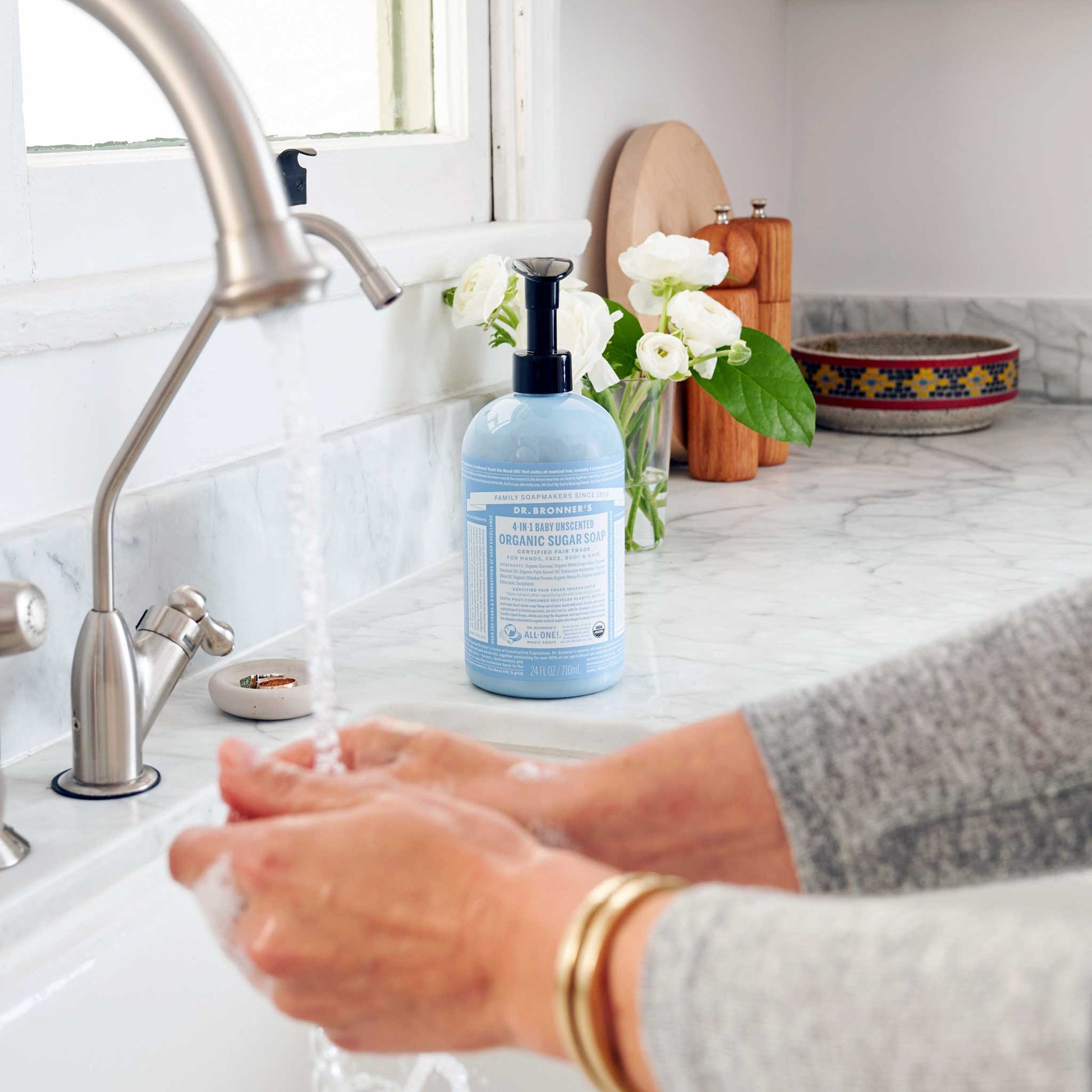 Hands lather with Dr. Bronners Organic Sugar Soap under a kitchen faucet, set against a bright countertop with decorative elements like flowers and dishes nearby [24 oz]