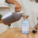 A person pours Dr Bronners 18 in 1 Baby Unscented Organic Sugar Soap from a large jug through a funnel into a smaller bottle on a wooden kitchen counter [12 oz]
