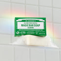 Dr. Bronners Magic Bar Soap in almond scent sits on a soap dish, generating bubbles against a tiled wall with a visible rainbow reflection. FAMILY SOAPMAKERS SINCE 1858, ALL ONE PURE CASTILE MAGIC BAR SOAP ALMOND, MADE WITH REGENERATIVE ORGANIC CERTIFIED OILS, FOR BODY FACE HANDS CERTIFIED FAIR TRADE [5 oz]