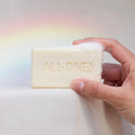 A hand holds a bar of soap with embossed text ALL ONE against a soft background with a visible rainbow. The scene suggests a natural, eco friendly product setting [5 oz]