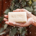 A hand holds a sudsy bar of soap with ALL ONE text embossed, against a blurred leafy background. This captures the essence of natural, vegan, eco friendly soap choices [5 oz]