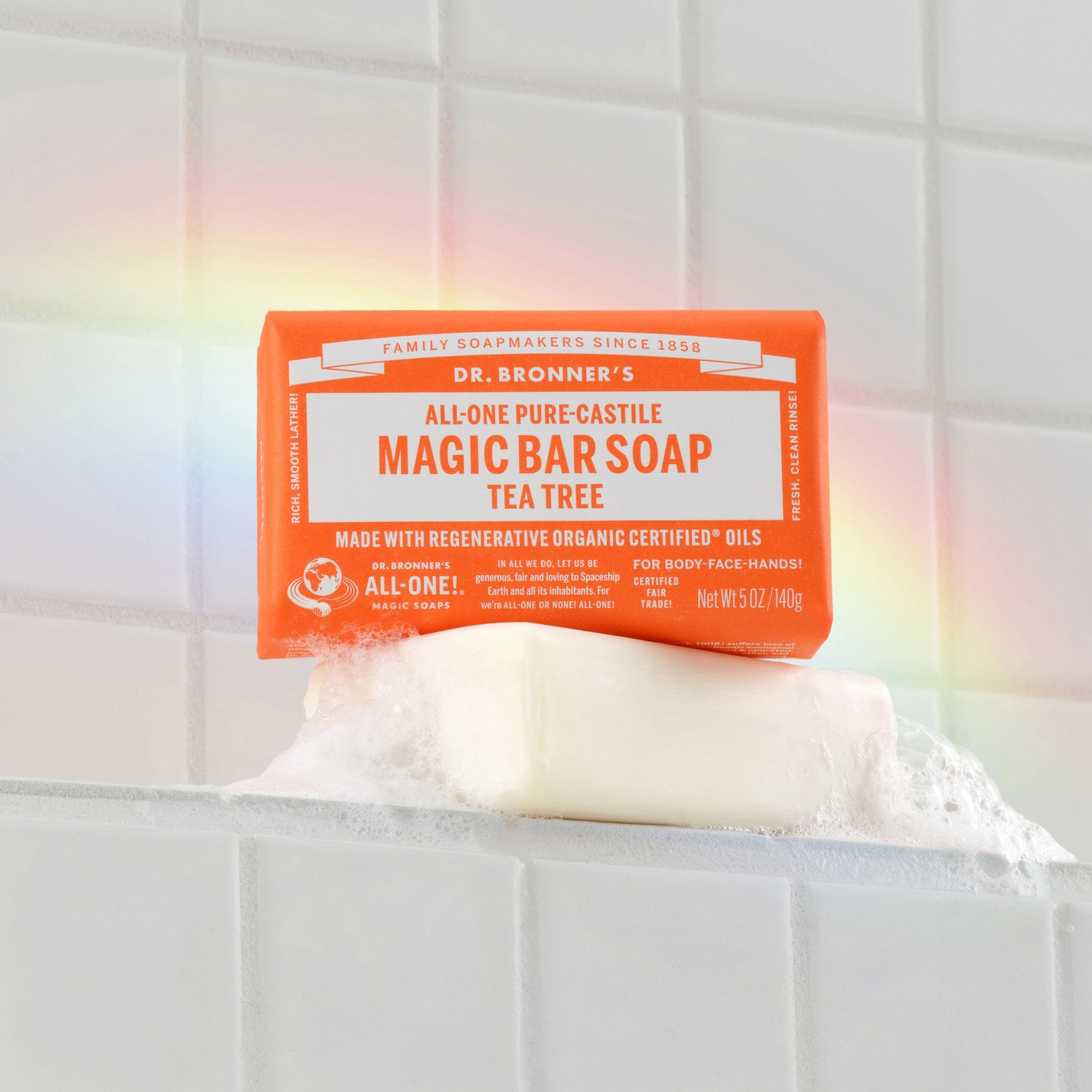 Bar soap rests on white tiled background, wrapped with an orange label. The text reads, Dr. Bronners All One Hemp Citrus Pure Castile Bar Soap. A subtle rainbow reflection illuminates the scene [5 oz]