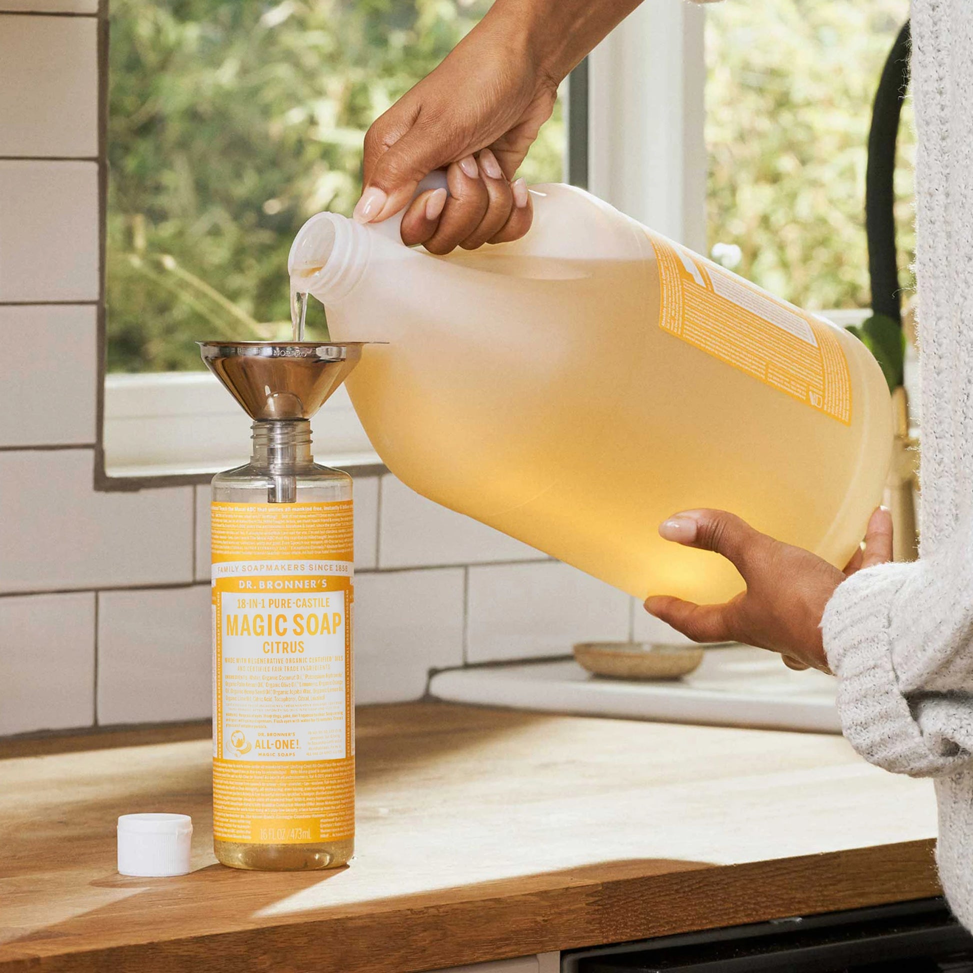 A large jug is pouring liquid soap into a smaller bottle via a funnel on a kitchen counter. The window behind suggests a well lit, natural setting [16 oz, 1 Gallon]
