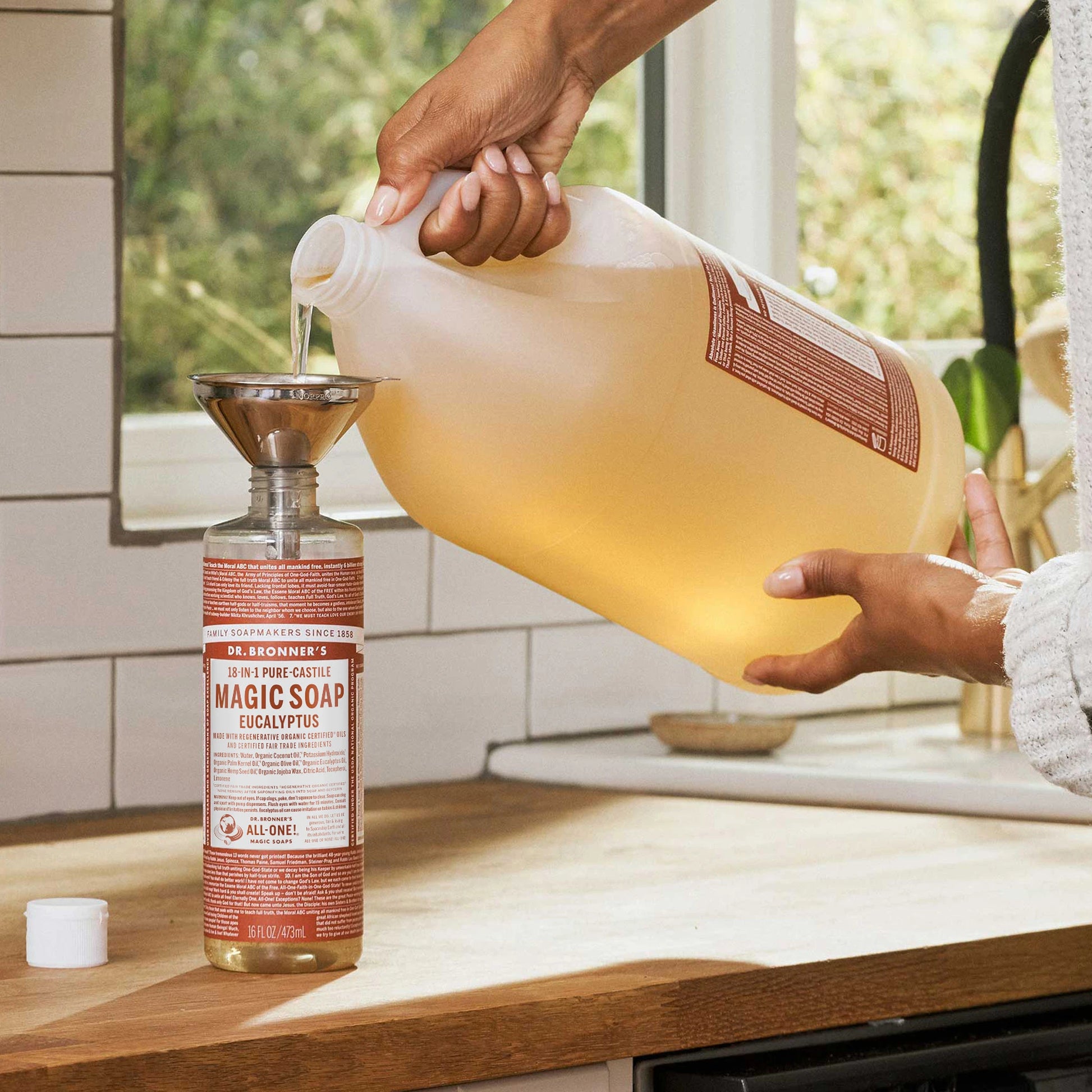 A person pours Dr. Bronners 18 in 1 Pure Castile Magic Soap Eucalyptus from a large container into a smaller bottle using a funnel. The scene is set in a kitchen with a wooden countertop [16 oz, 1 Gallon]