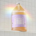 A large bottle of Dr. Bronners Lavender Pure Castile Soap sits on a tiled bathroom shelf, reflecting a rainbow light. The lavender label details its vegan, organic, and versatile cleaning uses [1 Gallon]