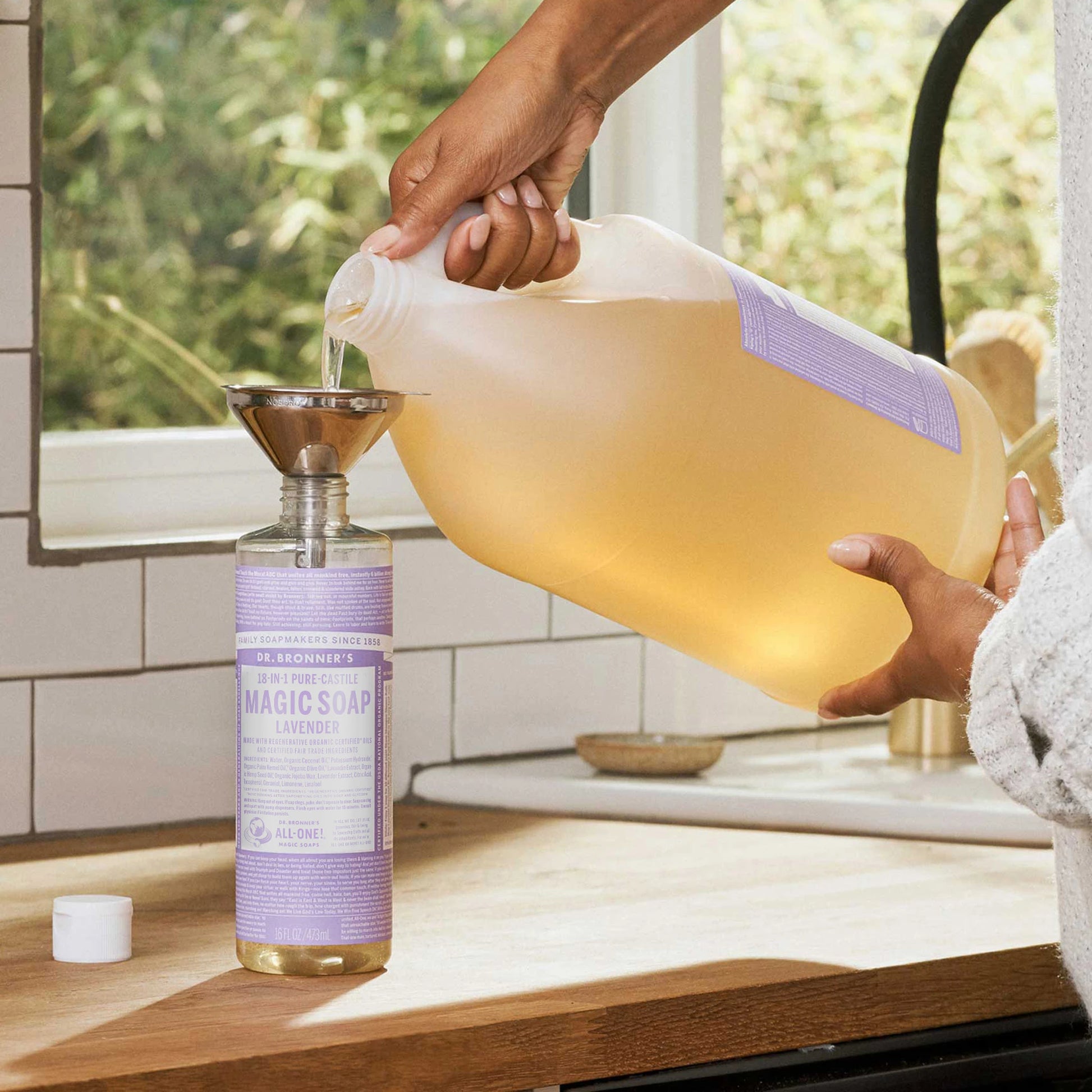 A lavender labeled bottle being refilled with liquid castile soap from a large jug using a funnel, set on a sunlit kitchen counter near a window overlooking greenery [16 oz]