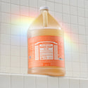 A gallon sized bottle of Dr. Bronners 18 in 1 Pure Castile Magic Soap in Tea Tree scent sits on a white tiled surface, framed by a subtle rainbow light effect [1 Gallon]
