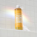 A bottle of Dr. Bronners 18 in 1 Pure Castile Magic Soap, Citrus, stands on a tiled ledge with soap suds. Rainbow light effects illuminate the scene, creating an eco friendly ambiance [16 oz]