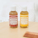 Two small bottles of Dr. Bronners liquid castile soap stand side by side on a wooden table. One is red labeled and the other yellow labeled. A comb lies nearby [2 oz]