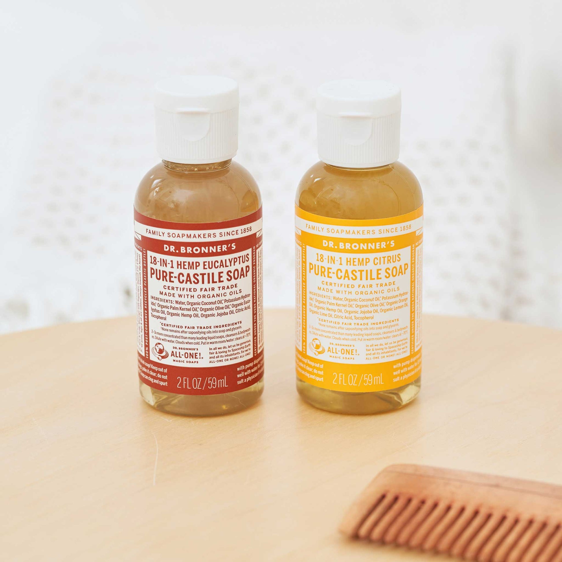 Two small bottles of Dr. Bronners liquid castile soap stand side by side on a wooden table. One is red labeled and the other yellow labeled. A comb lies nearby [2 oz]
