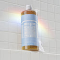 A bottle of Dr. Bronners Baby Unscented Pure Castile Liquid Soap stands vertically on a tiled surface, catching sunlight to create a subtle rainbow effect [32 oz]