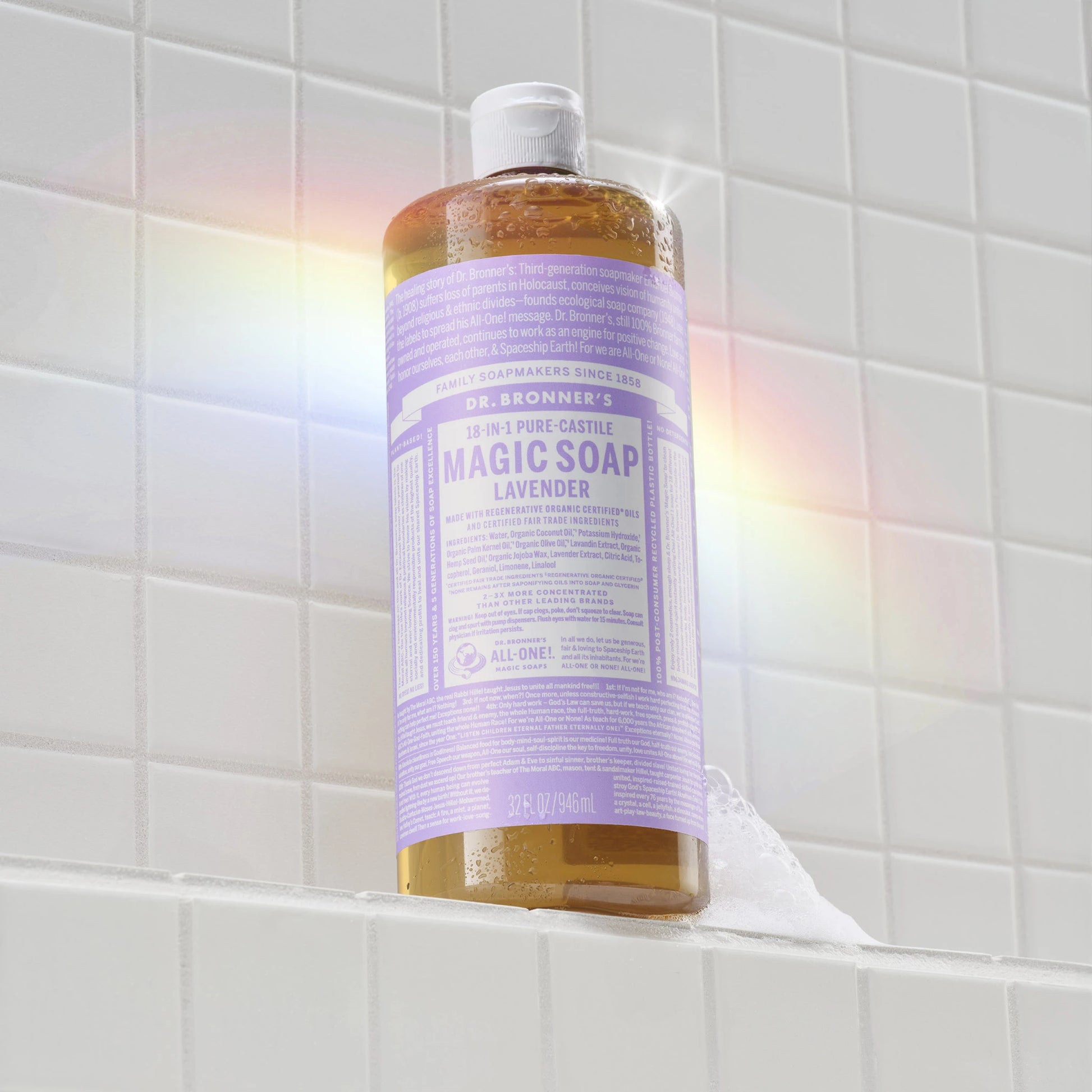 A bottle of Dr. Bronners lavender liquid castile soap stands on a white tiled shelf, illuminated by a soft rainbow reflection. Text on the bottle DR. BRONNERS 18 in 1 Pure Castile Soap [32 oz]