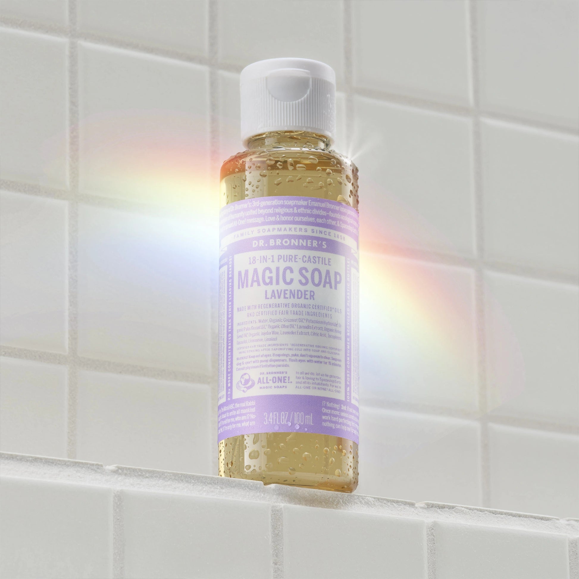 A small bottle of Dr. Bronners Pure Castile Magic Soap in lavender scent stands on a tiled surface, illuminated by a rainbow light. The label reads DR. BRONNERS 18 IN 1 PURE CASTILE MAGIC SOAP LAVENDER [3.4 oz]