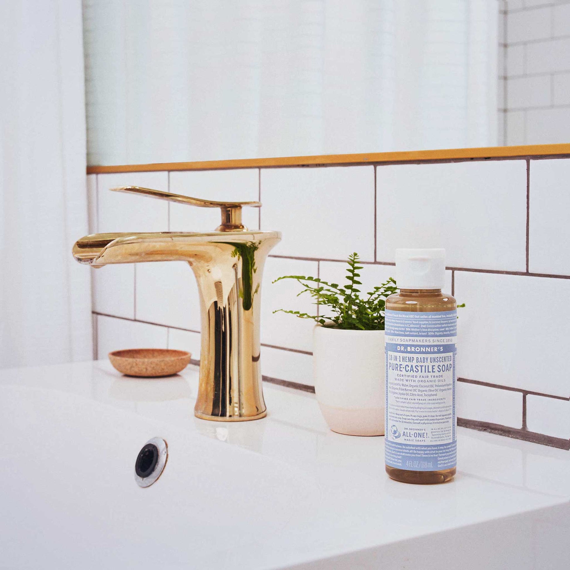 A bottle of Dr. Bronners Peppermint Pure Castile Soap sits on a white bathroom sink. A gold faucet and small plant enhance the clean, minimalist decor, highlighting sustainable soap use. Text on bottle DR. BRONNERS 18 IN 1 HEMP PEPPERMINT PURE CASTILE SOAP [4 oz]