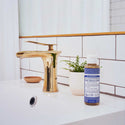 A bottle of Dr. Bronners 18 in 1 Hemp Peppermint Pure Castile Soap sits on a bathroom counter beside a gold faucet and a small potted plant, against tiled walls [4 oz]