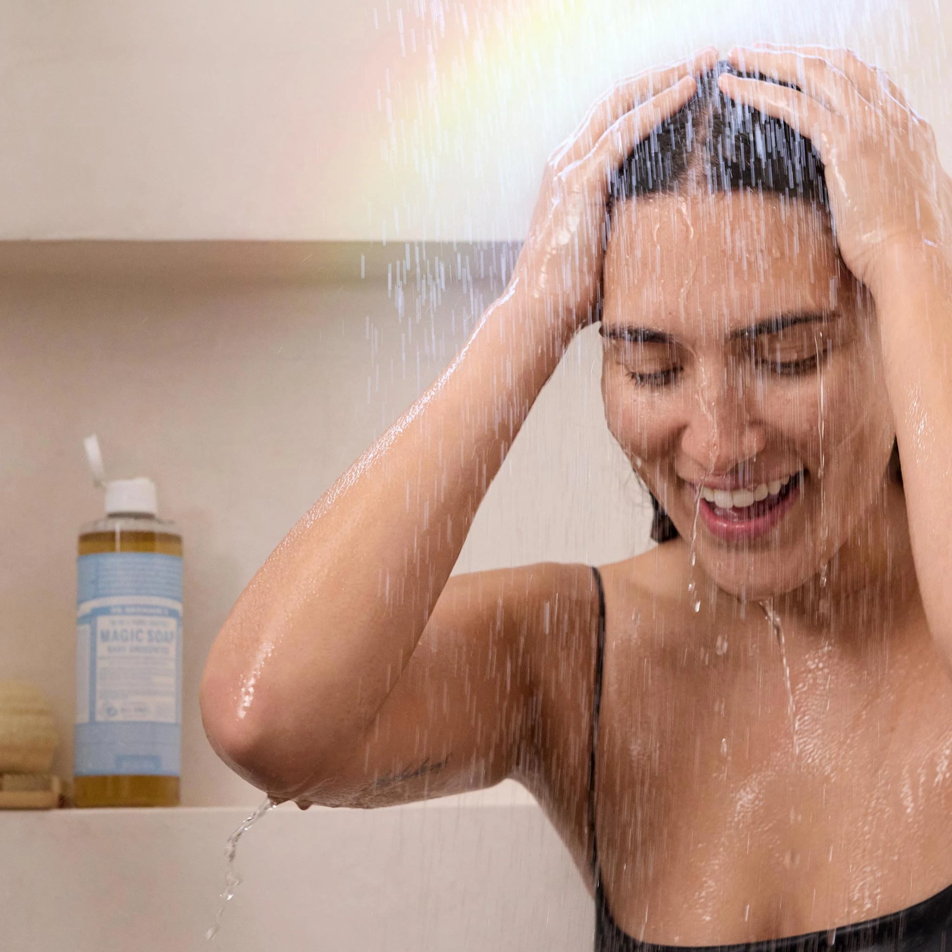 A person joyfully washes their hair under a shower. Behind them, a bottle of Dr. Bronners Pure Castile Soap, labeled 18 in 1 Hemp Peppermint, sits on a shelf. Streams of water create a refreshing ambiance [8 oz]
