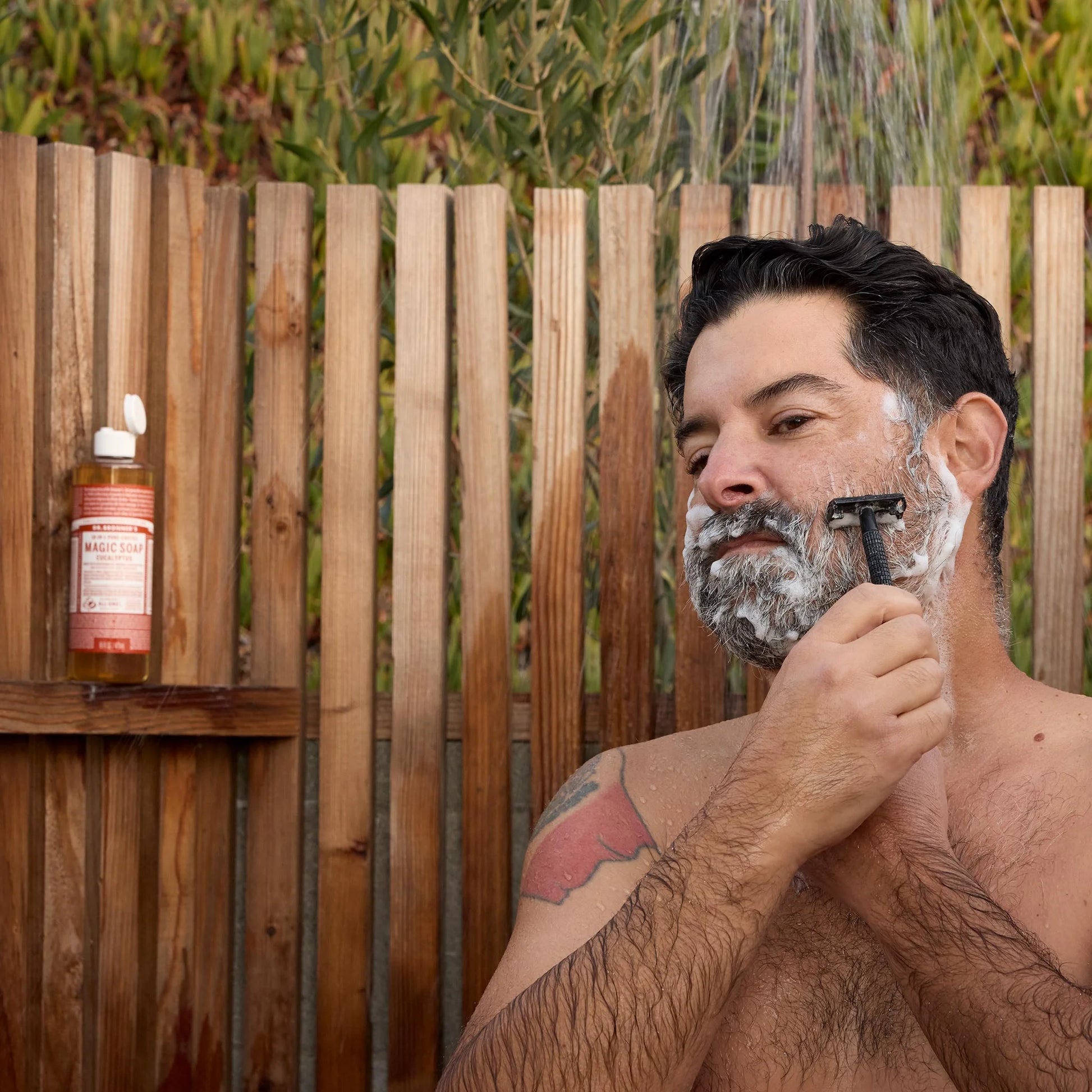 A man shaves outdoors using a razor and Dr Bronners soap, surrounded by a wooden fence and greenery. A bottle of Dr Bronners organic soap rests on a shelf nearby, enhancing the natural setting [8 oz]