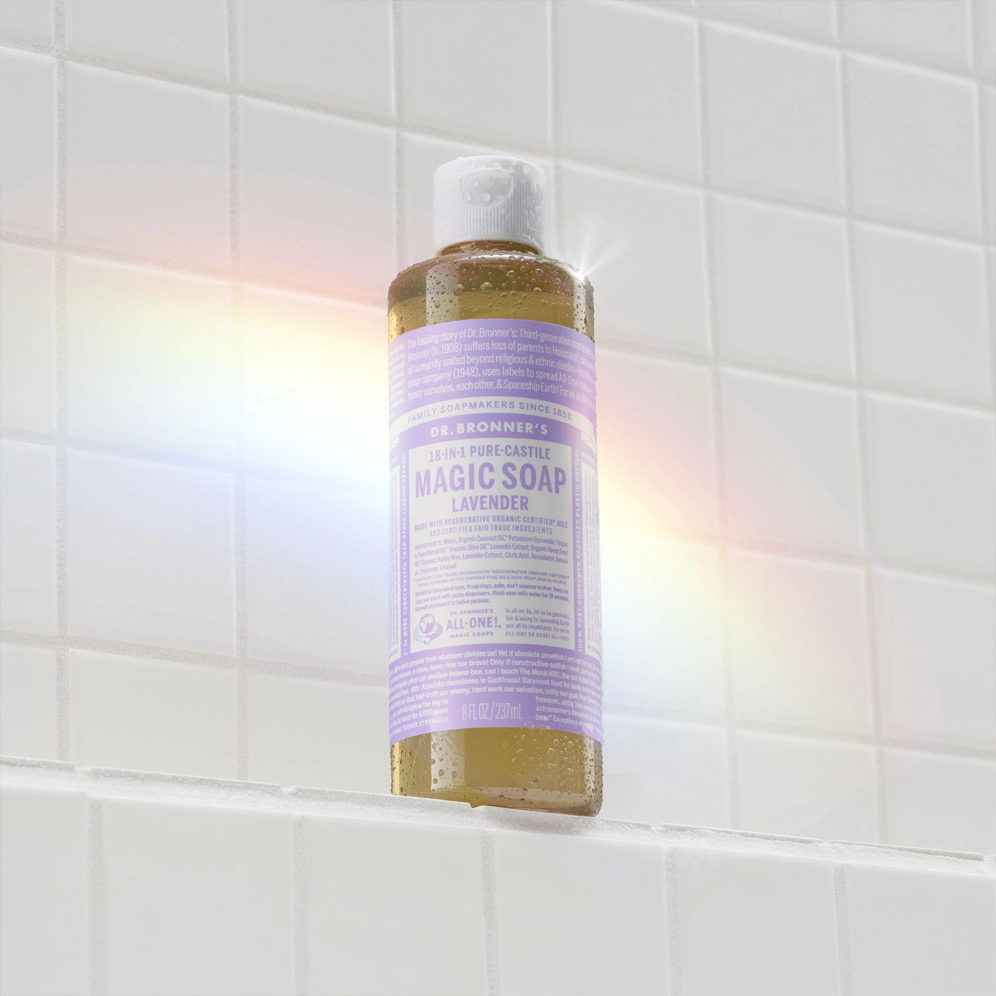 A bottle of Dr. Bronners 18 in 1 Pure Castile Lavender Magic Soap rests on a shower ledge. Text includes Family Soapmakers Since 1858, Made with Organic Oils, 8 fl. oz. . Bright, tiled bathroom backdrop [8 oz]
