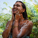 A person lathers organic castile soap on their face and body, smiling under a gentle outdoor shower. Surrounded by lush greenery, they enjoy a refreshing, eco friendly cleansing experience [2 oz, 3.4 oz, 4 oz, 8 oz, 16 oz, 32 oz, 1/2 Gallon, 1 Gallon]