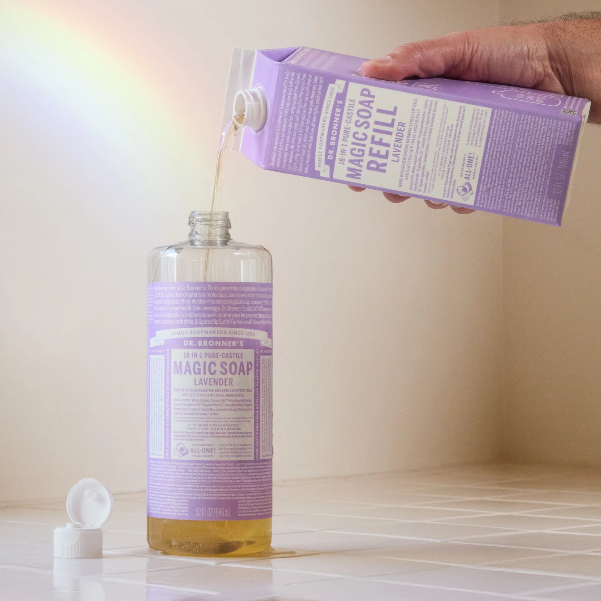 A hand pours Dr. Bronners lavender castile soap from a carton into a bottle on a white surface, with a cap nearby and a colorful light reflection on the wall [32 oz]