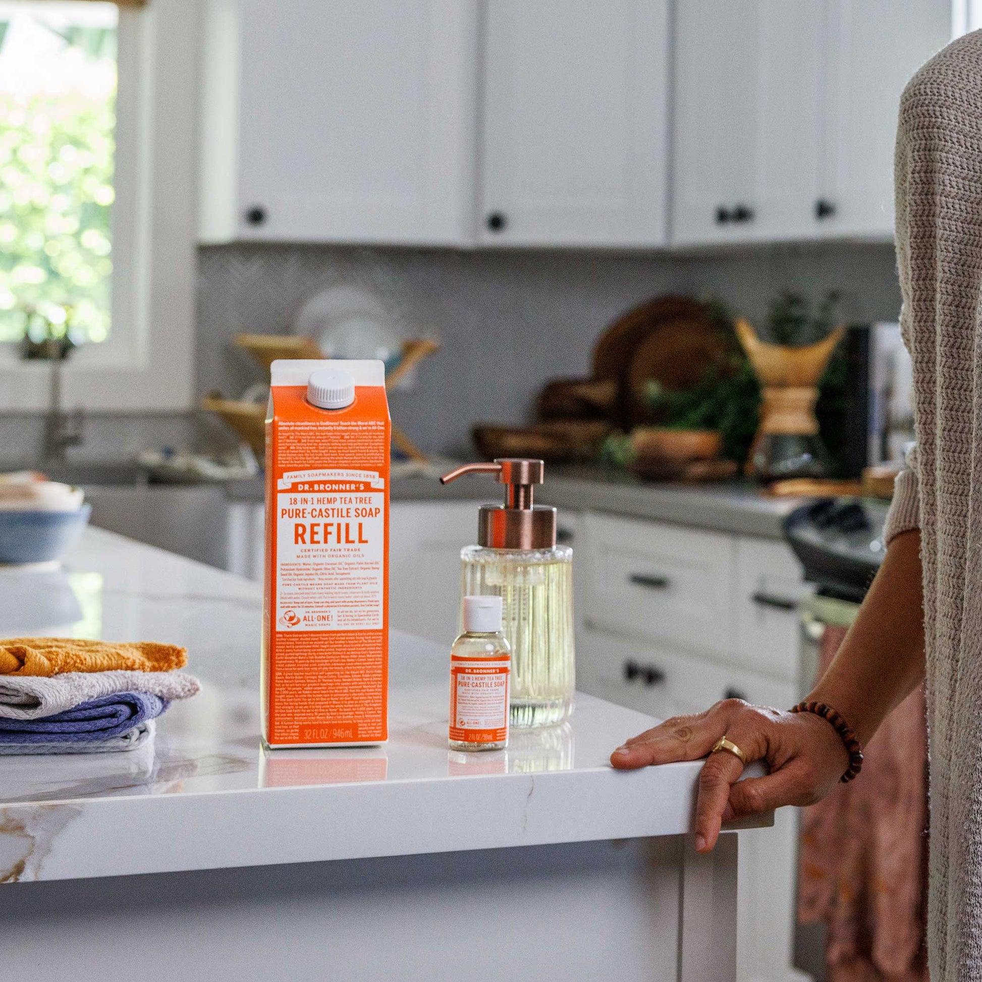 A bottle labeled Dr. Bronners and a liquid container sit on a kitchen countertop. A person stands nearby, with kitchen cabinets and a window in the background [32 oz]