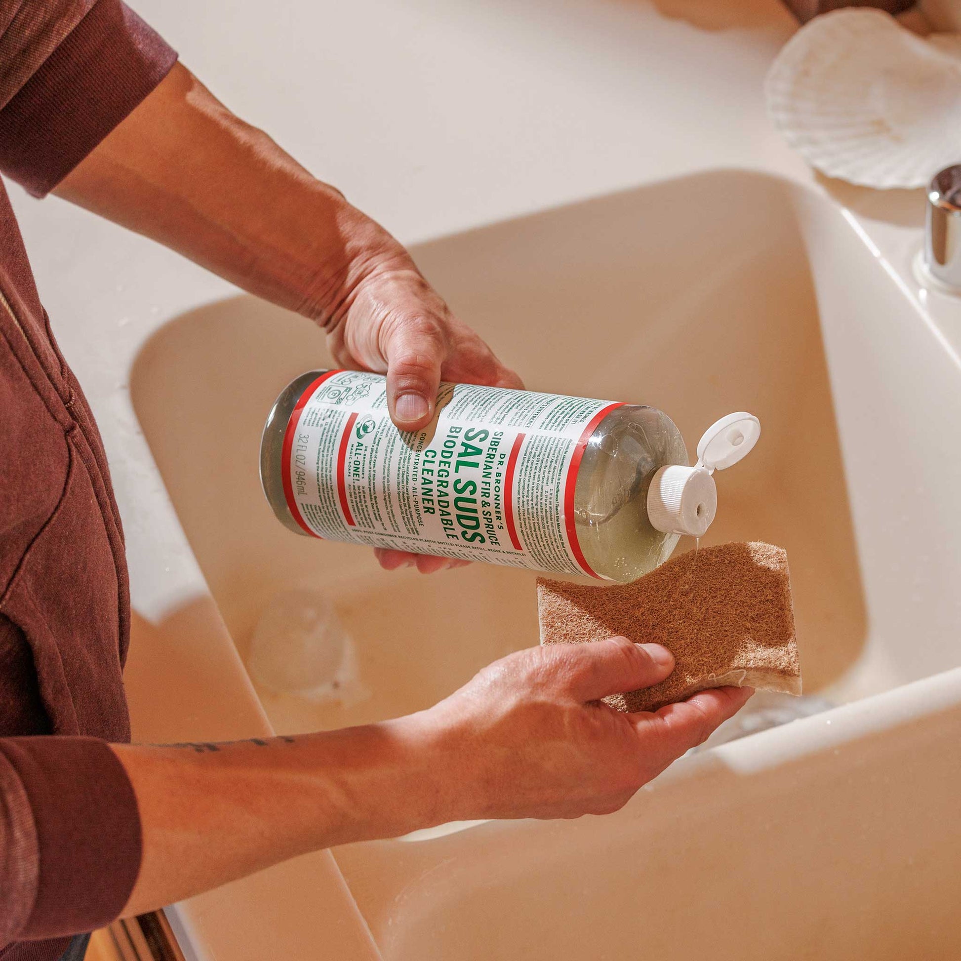 A person holds a bottle of Dr. Bronners peppermint Castile soap over a sink, preparing to use it. The environment appears to be a well lit bathroom with a countertop nearby [16 oz,32 oz]