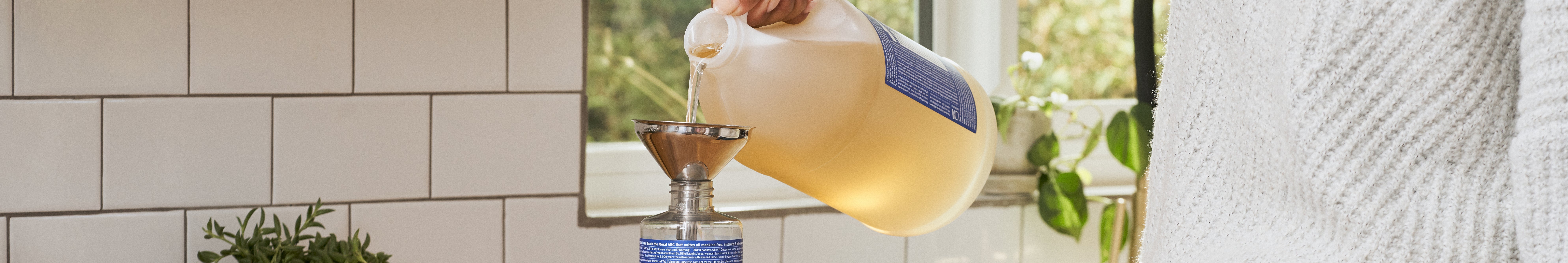 A person pours Dr. Bronners liquid castile soap from a large container into a funnel atop a smaller bottle in a tiled kitchen, emphasizing sustainable, eco friendly soap refilling practices