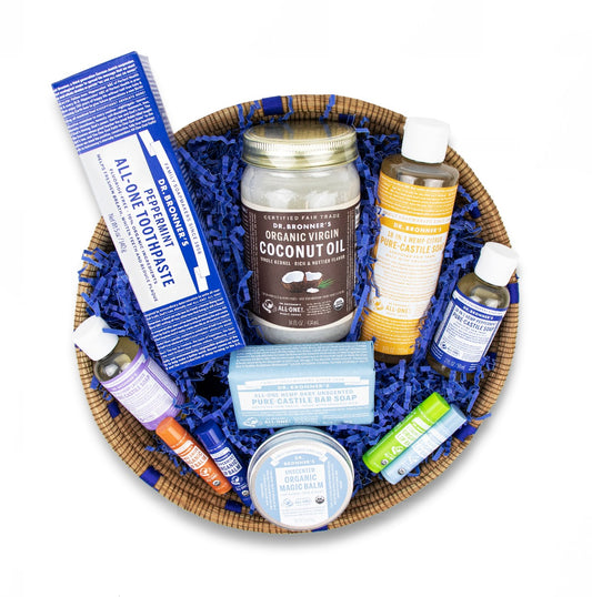 A basket contains various Dr. Bronners products, including organic virgin coconut oil, peppermint toothpaste, liquid Castile soaps, bar soap, and lip balms, nestled in blue decorative paper, ideal for eco friendly gift ideas