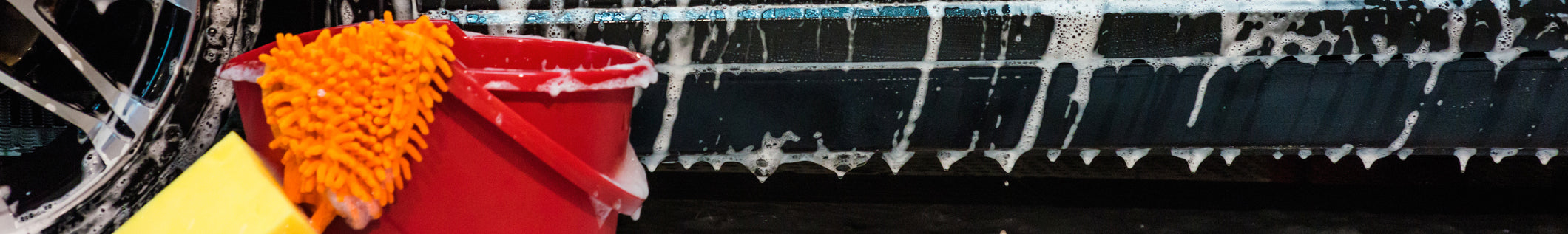 A red bucket filled with soapy water and an orange cleaning mitt rests beside a sponge, adjacent to a wet car rim and soapy car surface, ideal for eco friendly car cleaning