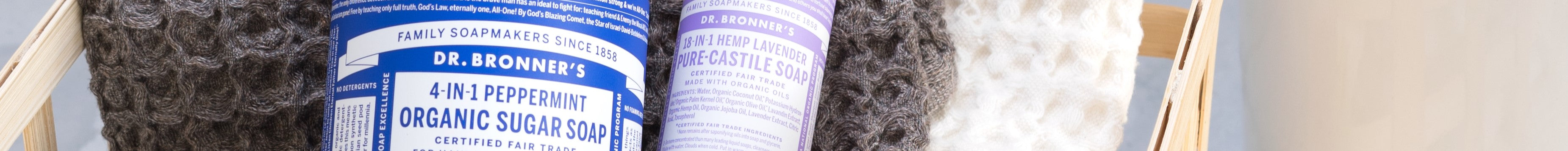 Bottles of Dr. Bronners soap, including 4 in 1 Peppermint Organic Sugar Soap and 18 in 1 Hemp Lavender Pure Castile Soap, rest on textured towels within a wooden bath caddy context
